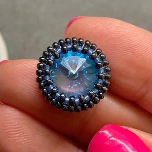 Könnte beinhalten: Ein rundes, mit Perlen besetztes Schmuckstück mit einem facettierten, hellblauen Mittelstein. Der Stein ist von einem Ring aus kleinen, dunkelblauen Perlen und einem zweiten Ring aus schwarzen Perlen umgeben. Das Schmuckstück wird in einer Hand mit pinkfarbenem Nagellack gehalten.