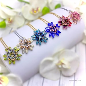 Puede incluir: Seis collares de flores con cuentas de varios colores, incluyendo verde, azul y rosa, expuestos sobre una superficie blanca. Cada collar presenta un colgante de flor de diferente color con una cadena delicada. Los collares están dispuestos en fila, mostrando la variedad de diseños.