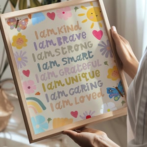 May include: A framed print with a light wood frame. The print features colorful text that reads "I am Kind, Brave, Strong, Smart, Grateful, Unique, Caring, Loved." The print is decorated with flowers, hearts, butterflies, a rainbow, and a smiling sun.