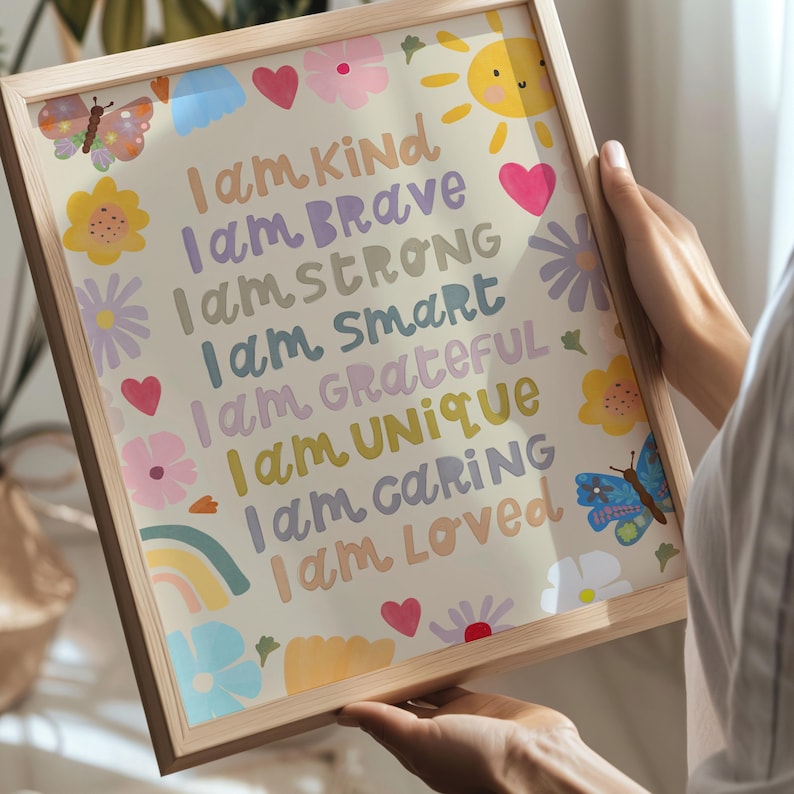 May include: A framed print with a light wood frame. The print features colorful text that reads "I am Kind, Brave, Strong, Smart, Grateful, Unique, Caring, Loved." The print is decorated with flowers, hearts, butterflies, a rainbow, and a smiling sun.