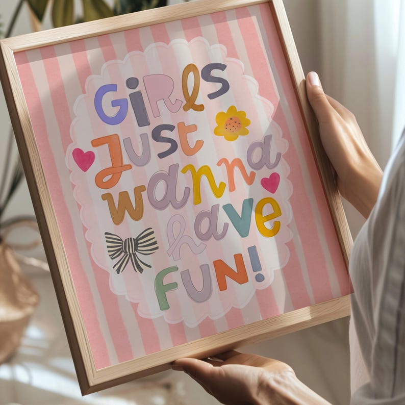 May include: A framed print with the words "Girls Just Wanna Have Fun" in colorful, playful lettering. The background features pink and white vertical stripes. The frame is made of light-colored wood.