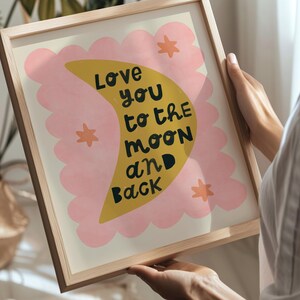 May include: A framed art print with a light wood frame. The artwork features a yellow crescent moon with the words "Love you to the moon and back" in black text. Pink cloud-like shapes and orange stars surround the moon.