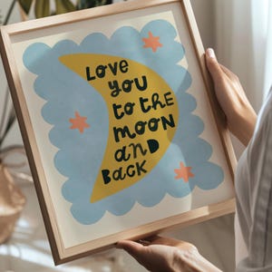 May include: A framed art print with the text "Love you to the moon and back." The artwork features a yellow crescent moon, blue cloud shapes, and orange stars on a cream background. The frame is made of light-colored wood.