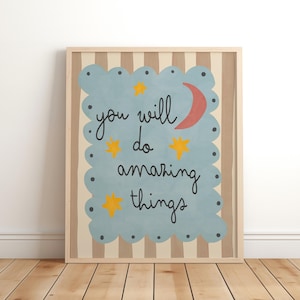 Puede incluir: Una impresión artística enmarcada con las palabras "you will do amazing things" en escritura negra. El texto está sobre un fondo azul claro en forma de nube con estrellas amarillas y una luna creciente roja. El fondo tiene un patrón de rayas beige y marrón.