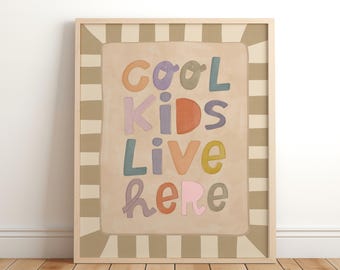 May include: A framed art print with the words "Cool Kids Live Here" in colorful, whimsical lettering. The text is set against a beige background with a striped border, all within a light wood frame.