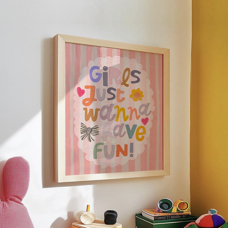 May include: A framed print with the text "Girls Just Wanna Have Fun!" in colorful letters. The background features pink and white vertical stripes. The frame is made of light-colored wood.