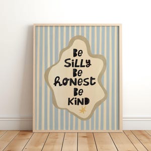 Op de afbeelding: Een ingelijste print met de woorden "Be Silly, Be Honest, Be Kind" in zwart op een beige, wolkvormig ontwerp. De achtergrond heeft verticale blauwe en crèmekleurige strepen. Het frame is lichtbruin.
