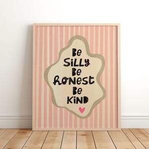Op de afbeelding: Een ingelijste kunstdruk met een licht houten frame. De achtergrond heeft verticale roze en crèmekleurige strepen. Het kunstwerk heeft een beige blob-vorm met de woorden "Be Silly Be Honest Be Kind" in zwart en een roze hart.