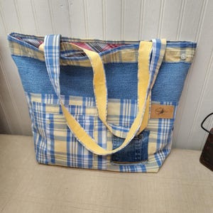 May include: A large tote bag with a patchwork design. The bag features denim, yellow, and blue plaid fabric, with yellow straps. A small denim pocket and a brown leather tag are visible on the front. The bag is designed for carrying items.