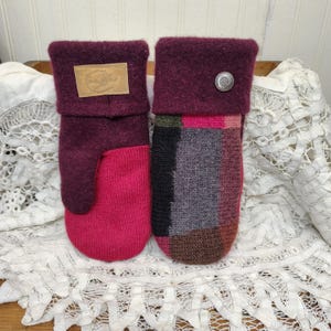 May include: A pair of handmade mittens in shades of burgundy and pink. One mitten features a solid burgundy cuff with a leather tag, while the other has a silver button. The lower portions are a patchwork of colors.