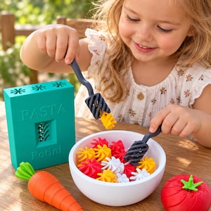 Può includere: Una scatola rettangolare color verde acqua per fare la pasta con la scritta PASTA Potini, una ciotola bianca riempita con piccoli pezzi di pasta a spirale gialli, rossi e bianchi, due forchette grigio scuro, una carota giocattolo arancione e un pomodoro rosso con gambo verde.