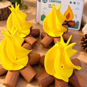 May include: Three miniature campfire figures featuring bright yellow plastic flames set atop brown textured log bases. Each piece is designed to resemble a small fire, and a game card labelled 13 CAMPFIRE is visible in the background.