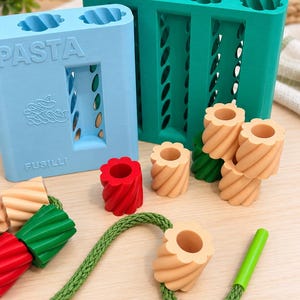 Może przedstawiać: Jasnoniebieska forma do makaronu z wytłoczonym napisem PASTA i FUSILLI z przodu, obok turkusowej formy. Kilka małych, cylindrycznych, spiralnych elementów przypominających makaron w kolorze czerwonym, zielonym i beżowym leży z zielonym sznurkiem.