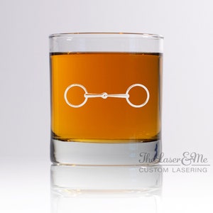 May include: A clear glass tumbler with a white etched design of a horse bit. The glass is filled with amber liquid and the bottom of the glass has the text "The Laser & Me CUSTOM LASERNG".