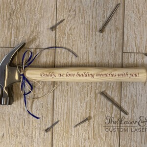 May include: A wooden hammer with a silver head, decorated with a blue ribbon and twine. The handle is engraved with the text "Daddy, we love building memories with you!"