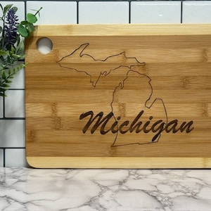 May include: A natural bamboo cutting board with a laser-etched outline of the state of Michigan and the word "Michigan" in a cursive font.