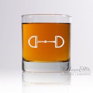 May include: A clear glass tumbler with a brown liquid inside. The glass has a white engraved design of a horse bit on the front. The text "The Laser & Me CUSTOM LASERNG" is on the bottom of the glass.