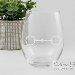 May include: A clear glass wine glass with a white engraved horse bit design. The glass is sitting on a white surface. The text "The Laser & Me CUSTOM LASER" is visible at the bottom of the image.