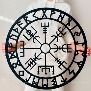 May include: A black wooden wall decor featuring a circular compass with a black and white design. The compass is surrounded by a circle of runes.