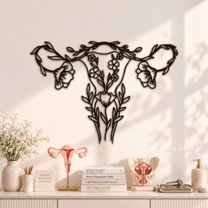 May include: A black metal wall art piece shaped like a human uterus, featuring intricate floral and vine motifs integrated into the fallopian tubes and ovaries. The design includes stylized flowers and leaves throughout the structure.