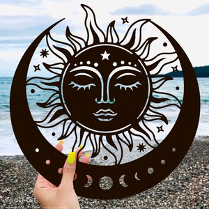 May include: Black and white sun and moon design with a face on the sun. The design is cut out of wood and is being held in front of a beach scene.