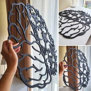 May include: A black laser-cut wooden wall art piece shaped like a human brain. One half of the brain features intricate, branching tree-like patterns, while the other half is composed of detailed, symmetrical floral and botanical cutouts.