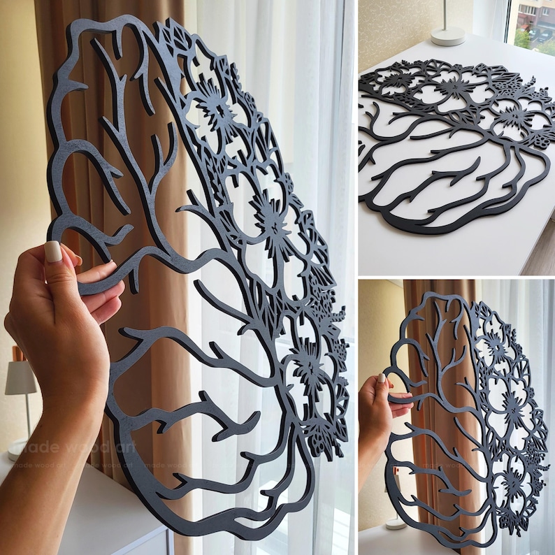 May include: A black laser-cut wooden wall art piece shaped like a human brain. One half of the brain features intricate, branching tree-like patterns, while the other half is composed of detailed, symmetrical floral and botanical cutouts.
