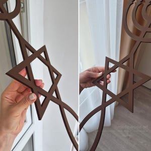 May include: Brown wooden Star of David wall art. The six-pointed star is a prominent feature, with a decorative menorah design. The piece is held up in front of a window and a curtain.