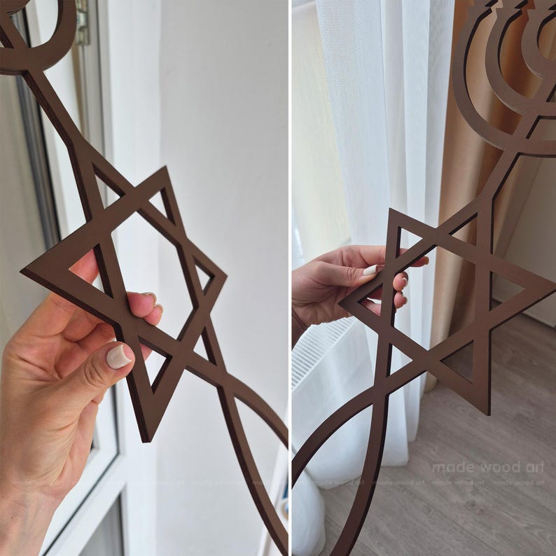 May include: Brown wooden Star of David wall art. The six-pointed star is a prominent feature, with a decorative menorah design. The piece is held up in front of a window and a curtain.