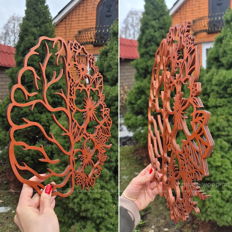 May include: A laser-cut wooden wall art piece featuring a split design. One side depicts the anatomical structure of a human brain with intricate sulci and gyri, while the other side displays a detailed floral pattern with various blossoms.