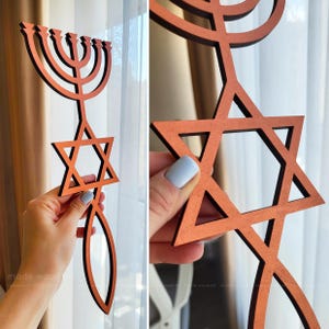 May include: A wooden, orange-colored Star of David and menorah. The Star of David is in the center, with a menorah at the top and a stylized fish at the bottom. The menorah has nine candle holders. The item is made of wood.