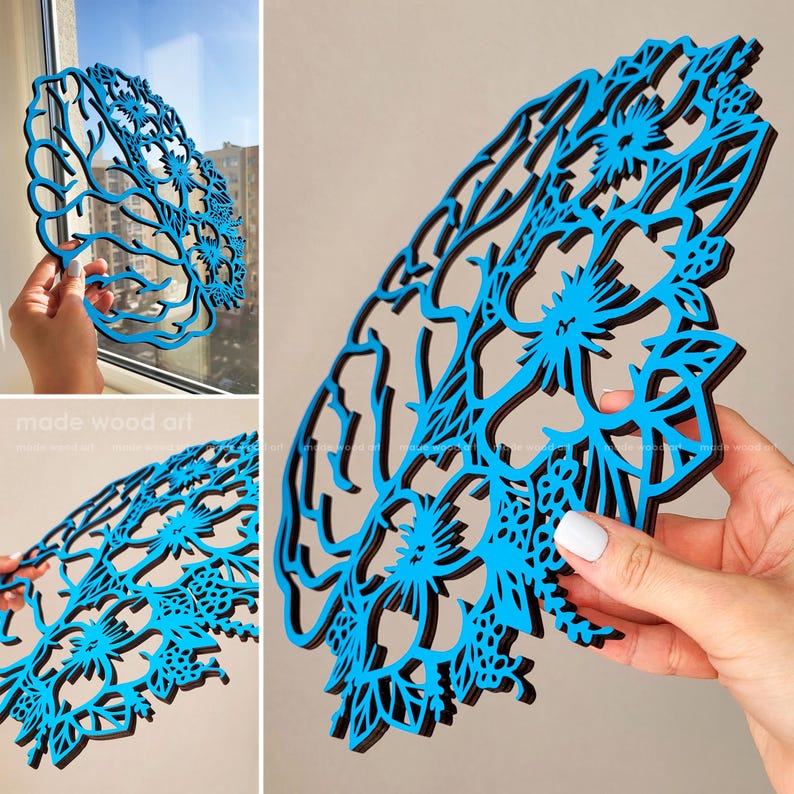 May include: A laser-cut wooden wall art piece shaped like a human brain, featuring a bright blue front layer with intricate floral and leaf cutouts layered over a solid black base, creating a detailed anatomical silhouette with decorative patterns.