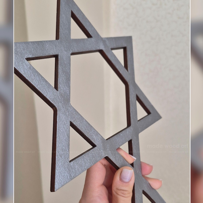 May include: A gray wooden Star of David wall hanging. The six-pointed star is cut out of wood, with a dark brown edge. The design is held by a person's hand. The background is a light beige wall.