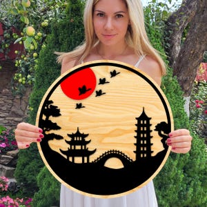 May include: A round wooden wall art piece with a black silhouette of a Japanese garden scene. The scene includes a pagoda, a bridge, trees, and birds flying over a red sun.