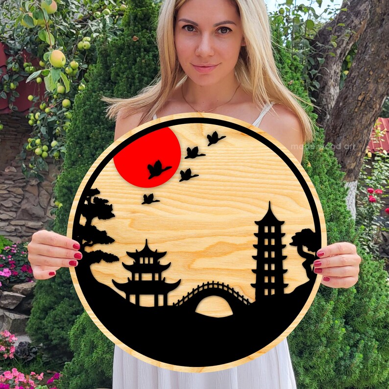 May include: A round wooden wall art piece with a black silhouette of a Japanese garden scene. The scene includes a pagoda, a bridge, trees, and birds flying over a red sun.