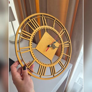 May include: A wooden wall clock with a gold finish. The clock features Roman numerals and a square center. The hands are black with a red second hand. The clock is held by a person.