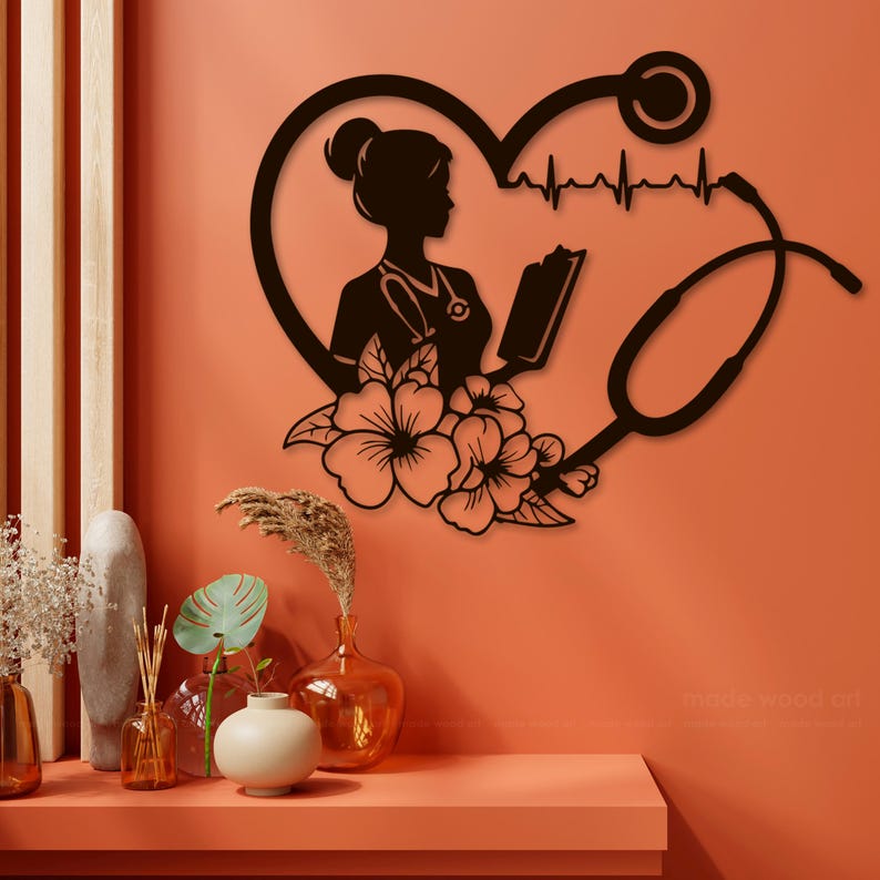 Op de afbeelding: Zwarte metalen wandkunst met een silhouet van een verpleegster die een boek, een stethoscoop en een hart met een hartslaglijn vasthoudt. Het ontwerp is versierd met bloemmotieven.