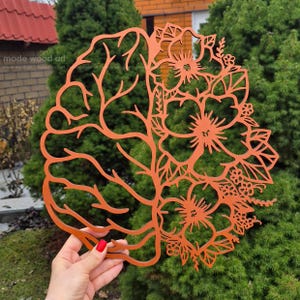 May include: A laser-cut orange wooden wall art piece featuring a split design. The left side depicts the anatomical structure of a human brain with intricate branching lines, while the right side displays a collection of detailed floral blossoms.