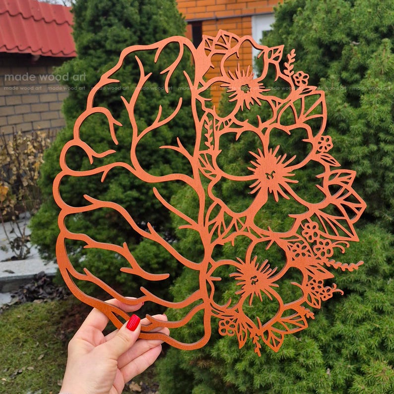 May include: A laser-cut orange wooden wall art piece featuring a split design. The left side depicts the anatomical structure of a human brain with intricate branching lines, while the right side displays a collection of detailed floral blossoms.