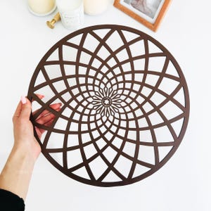 May include: A brown wooden round placemat with a geometric design. The design is a spiral of overlapping squares.
