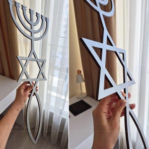 May include: Wooden wall art featuring a Menorah, Star of David, and a fish symbol. The art piece is made of dark brown wood with a light gray interior. The Menorah has nine candle holders. The Star of David is a six-pointed star.