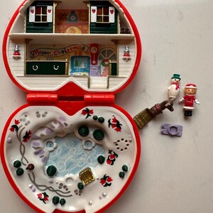 Può includere: Un compatto Polly Pocket vintage a tema natalizio a forma di cuore, aperto per rivelare una scena di casa in miniatura. Il compatto è rosso e bianco con interni dettagliati, tra cui piccole figure di un pupazzo di neve e una ragazza con un cappotto rosso.