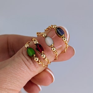 May include: Four gold-colored wire rings, each with a different colored oval bead: green, red, white, and dark blue. The rings are held up by a finger, showcasing the jewelry's design and colors.