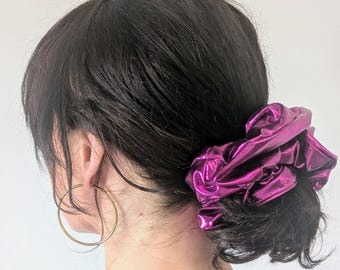 May include: A metallic magenta scrunchie made of a shiny, reflective fabric gathered around dark hair. The hair accessory is paired with a thin gold hoop earring and a gold chain necklace, visible against a plain, light-colored background.