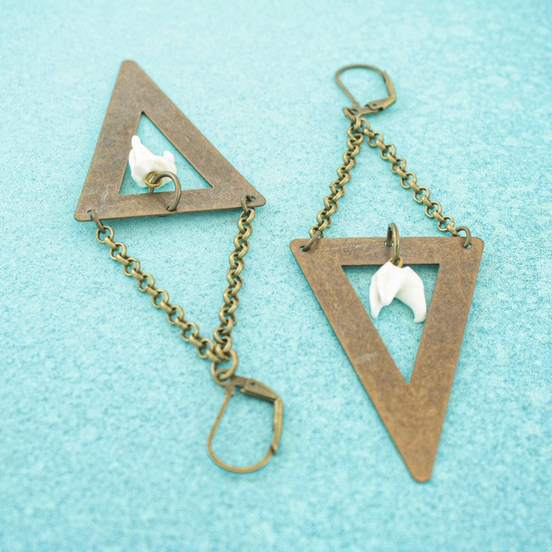 May include: A pair of geometric earrings with a triangular frame in a bronze tone. Each earring features a white, organic-shaped charm suspended within the triangle and connected by a chain. The earrings have a lever-back closure.