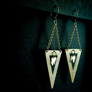 May include: A pair of unique earrings featuring a triangular brass pendant with a small chain and a dangling tooth. The earrings have a dark background and are suspended from a black bar.