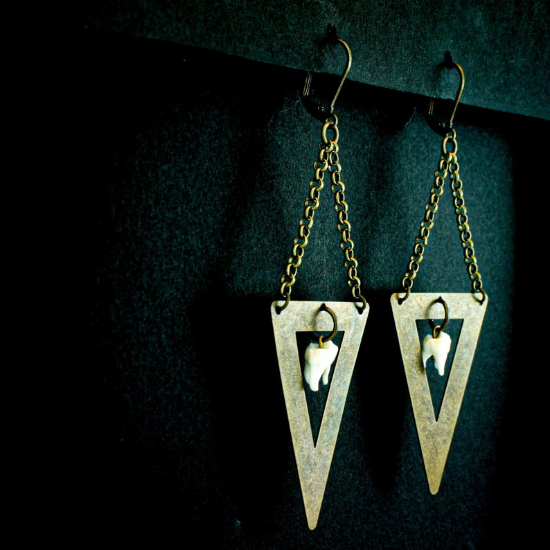 May include: A pair of unique earrings featuring a triangular brass pendant with a small chain and a dangling tooth. The earrings have a dark background and are suspended from a black bar.