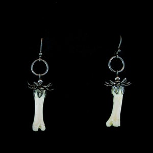 Puede incluir: Un par de pendientes únicos con colgantes en forma de hueso. Cada pendiente tiene un anillo de metal plateado y un elemento decorativo en la parte superior del hueso. El fondo es negro liso.