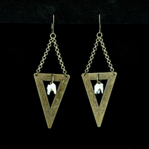May include: A pair of unique earrings featuring a brass-colored triangle design. Each earring has a small, white tooth suspended within the triangle, connected by a delicate chain. The earrings hang from a hook.