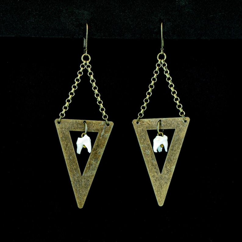 May include: A pair of unique earrings featuring a brass-colored triangle design. Each earring has a small, white tooth suspended within the triangle, connected by a delicate chain. The earrings hang from a hook.